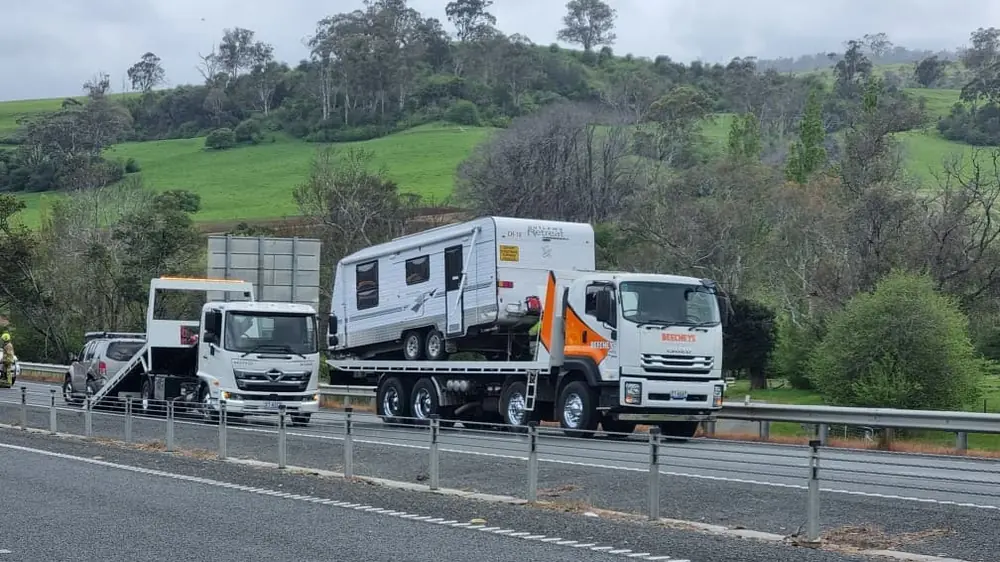 A Beecheys Towing flatbed carrying a caravan past green hills on the highway