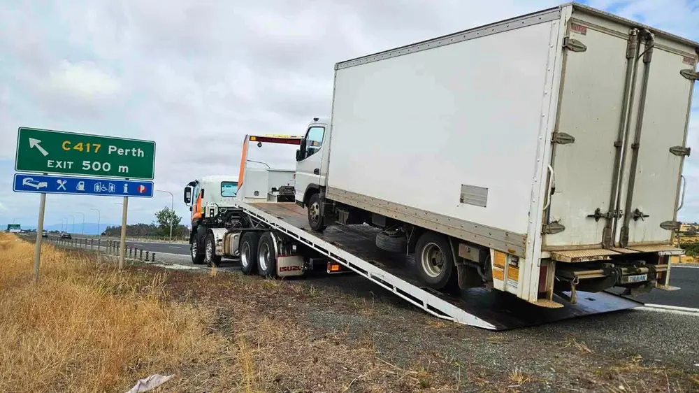 A Beecheys Towing flatbed recovering a box truck on the highway shoulder near a Perth exit sign