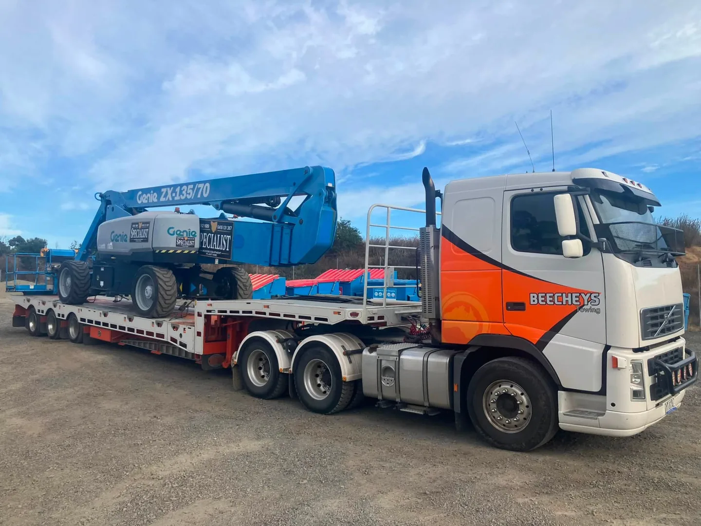 Beecheys Towing low-loader transporting a blue Genie ZX-135/70 boom lift on a gravel yard