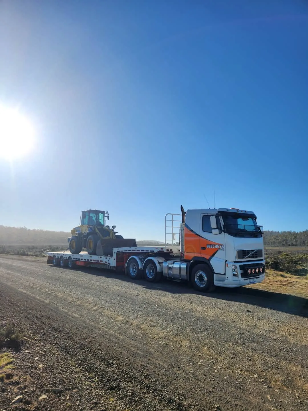 Beecheys Towing Volvo prime mover and low-loader trailer carrying a Komatsu wheel loader on a gravel road in the Tasmanian highlands